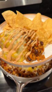 A close-up of a plate with a cheesy taco casserole slice topped with a spicy cream drizzle, served with tortilla chips, sitting above the pot it was scooped from.