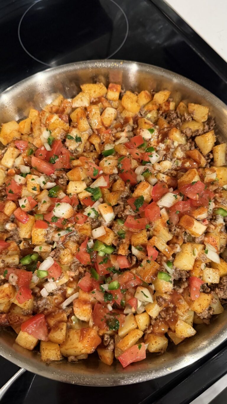An overhead view of a skillet filled with seasoned diced potatoes, ground beef, and fresh pico de gallo-style toppings.