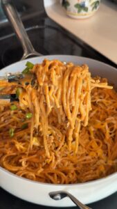 A pair of tongs lifts a tangle of saucy noodles from a skillet filled with a noodle dish garnished with bean sprouts and green onions.