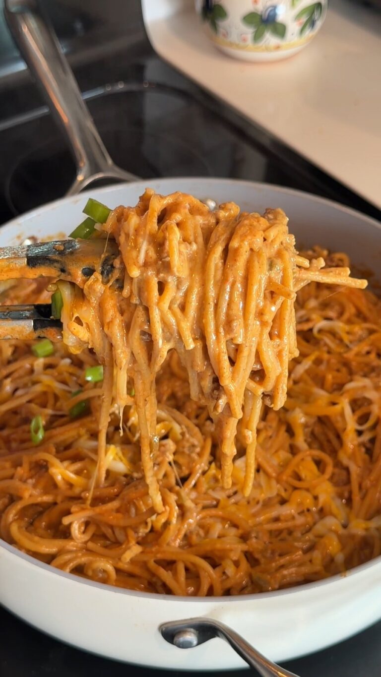 A pair of tongs lifts a tangle of saucy noodles from a skillet filled with a noodle dish garnished with bean sprouts and green onions.