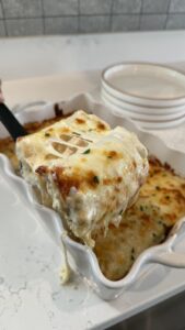 A close-up of a cheesy, golden-baked lasagna slice being lifted from a white baking dish with a spatula.