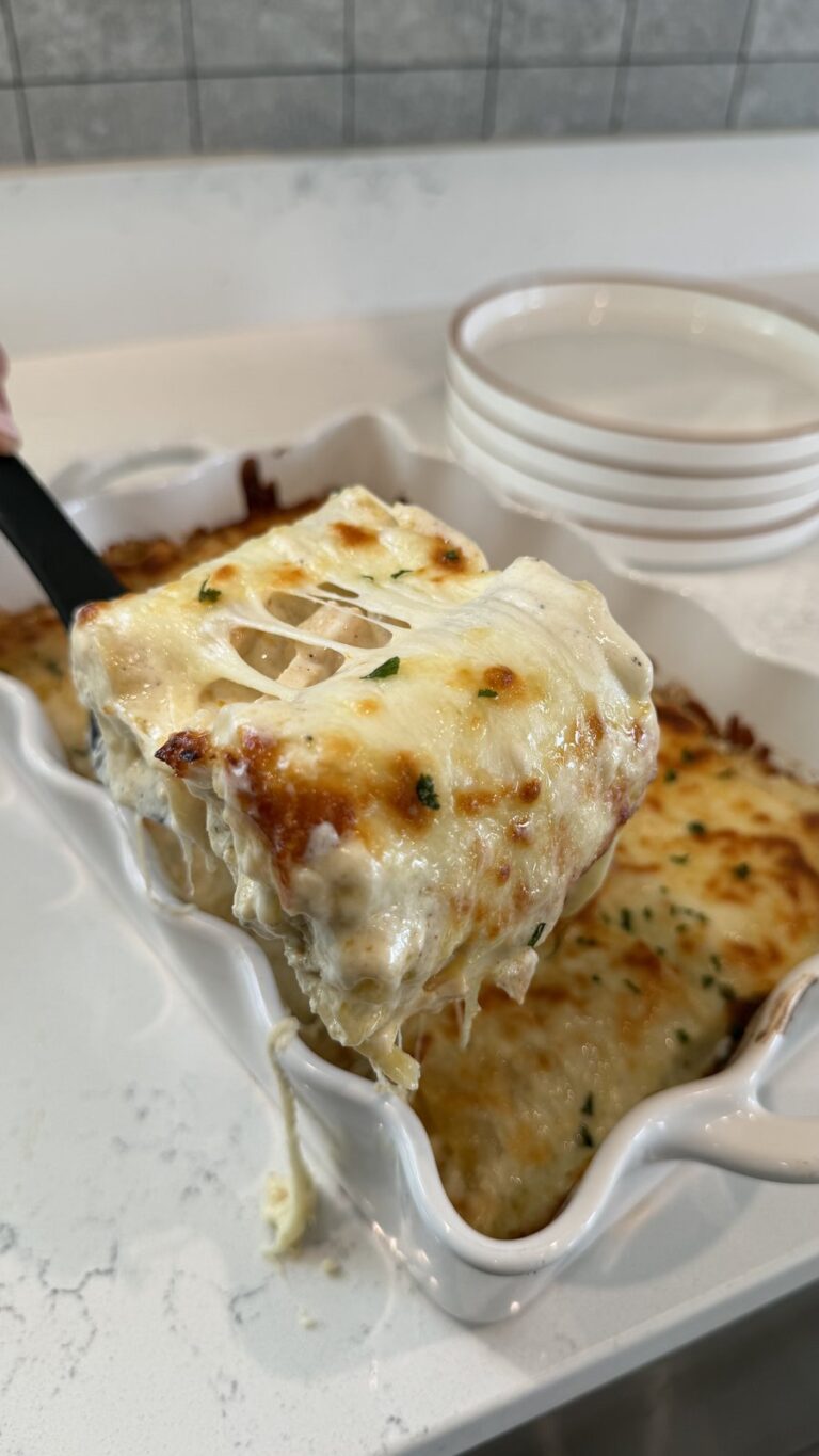 A close-up of a cheesy, golden-baked lasagna slice being lifted from a white baking dish with a spatula.