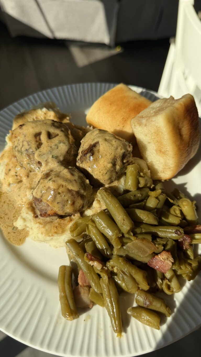A plate of Swedish-style meatballs in gravy over mashed potatoes, served with dinner rolls and seasoned green beans.