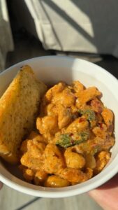 A hand holds a white bowl containing creamy tomato chicken pasta with spinach alongside a slice of garlic bread.