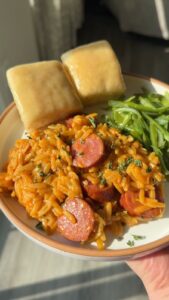 A plate of orange rice with sliced sausage, green beans, and bread rolls held by a person's hand.