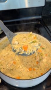 A ladle lifts creamy chicken noodle soup with carrots and celery from a pot on a stove.