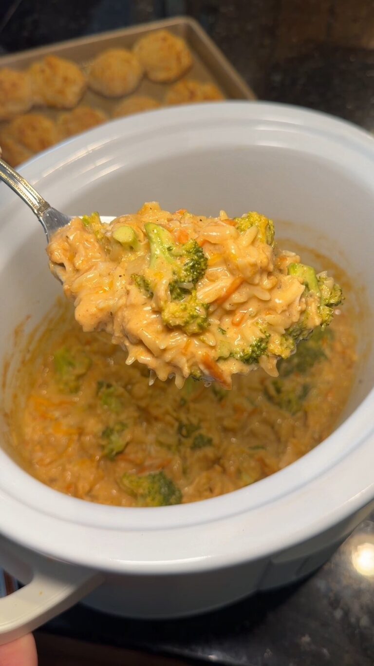 A spoon lifts a serving of creamy cheesy broccoli and rice casserole from a slow cooker, with a tray of chicken nuggets in the background.