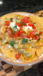 A plate of loaded nachos topped with melted cheese, chicken, sour cream, and fresh pico de gallo sits on a wooden board.