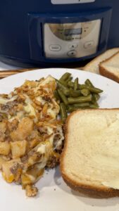 A plate of cheesy meat and potato casserole with green beans and buttered bread, served in front of a slow cooker.