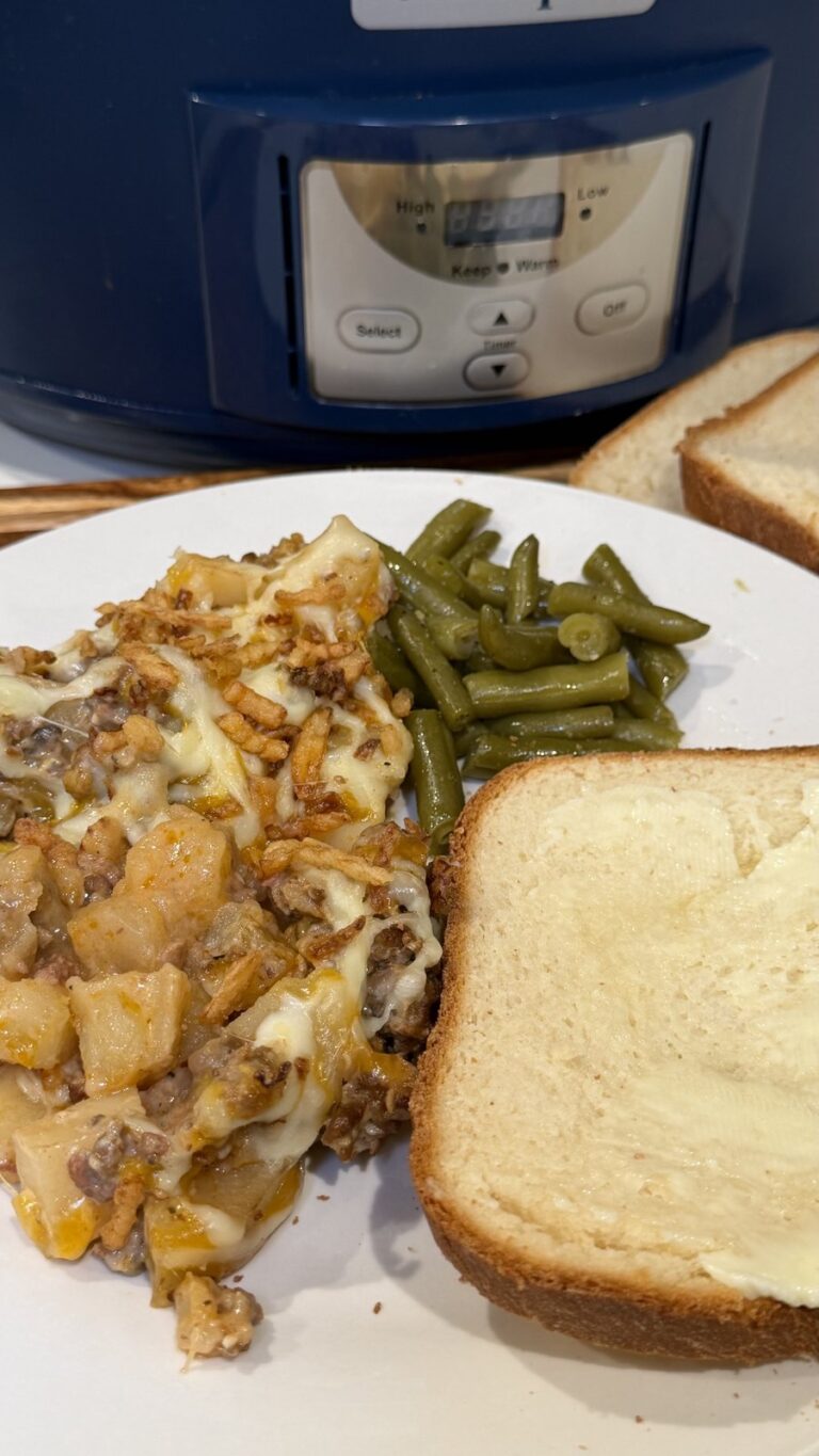 A plate of cheesy meat and potato casserole with green beans and buttered bread, served in front of a slow cooker.