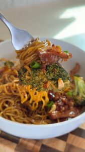 A close-up shot of a fork lifting spicy noodles topped with broccoli, beef, and chili flakes from a white bowl.