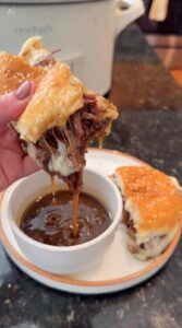 A hand dips a cheesy shredded beef sandwich into a bowl of au jus with another sandwich half and a crockpot visible in the background.