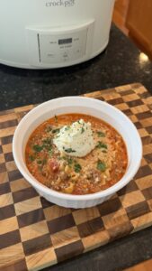 A bowl of lasagna soup topped with a dollop of ricotta cheese, parmesan, and fresh herbs sits on a wooden cutting board in front of a crock-pot slow cooker.