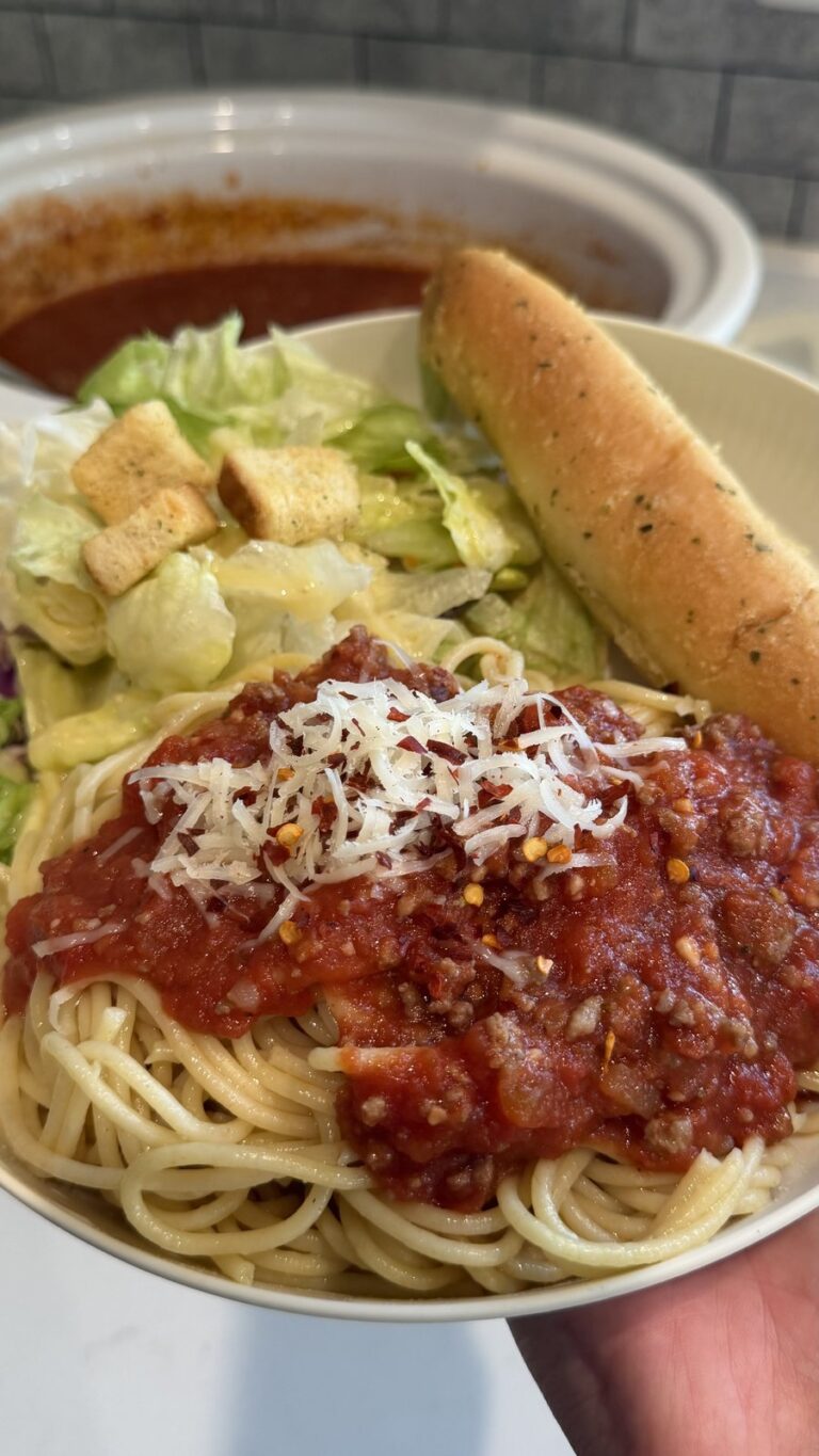 A hand holds a bowl of spaghetti topped with meat sauce and parmesan, alongside a caesar salad and a breadstick.