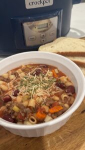 A bowl of hearty pasta e fagioli soup with beans, ground beef, and vegetables, topped with parmesan, sits next to slices of bread in front of a crockpot.