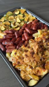 An overhead view of a sheet pan filled with roasted zucchini and onions, cocktail hot dogs, and cheesy bacon-topped potato wedges.