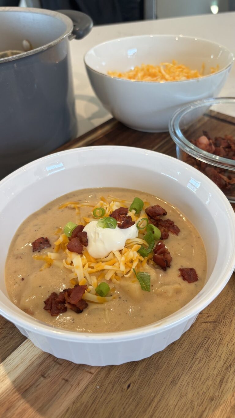 A bowl of creamy potato soup topped with shredded cheese, bacon bits, sour cream, and green onions, with additional toppings in bowls nearby.