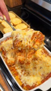 A hand lifts a cheesy scoop of baked ziti pasta with a spoon from a casserole dish, with garlic bread toasts in the background.