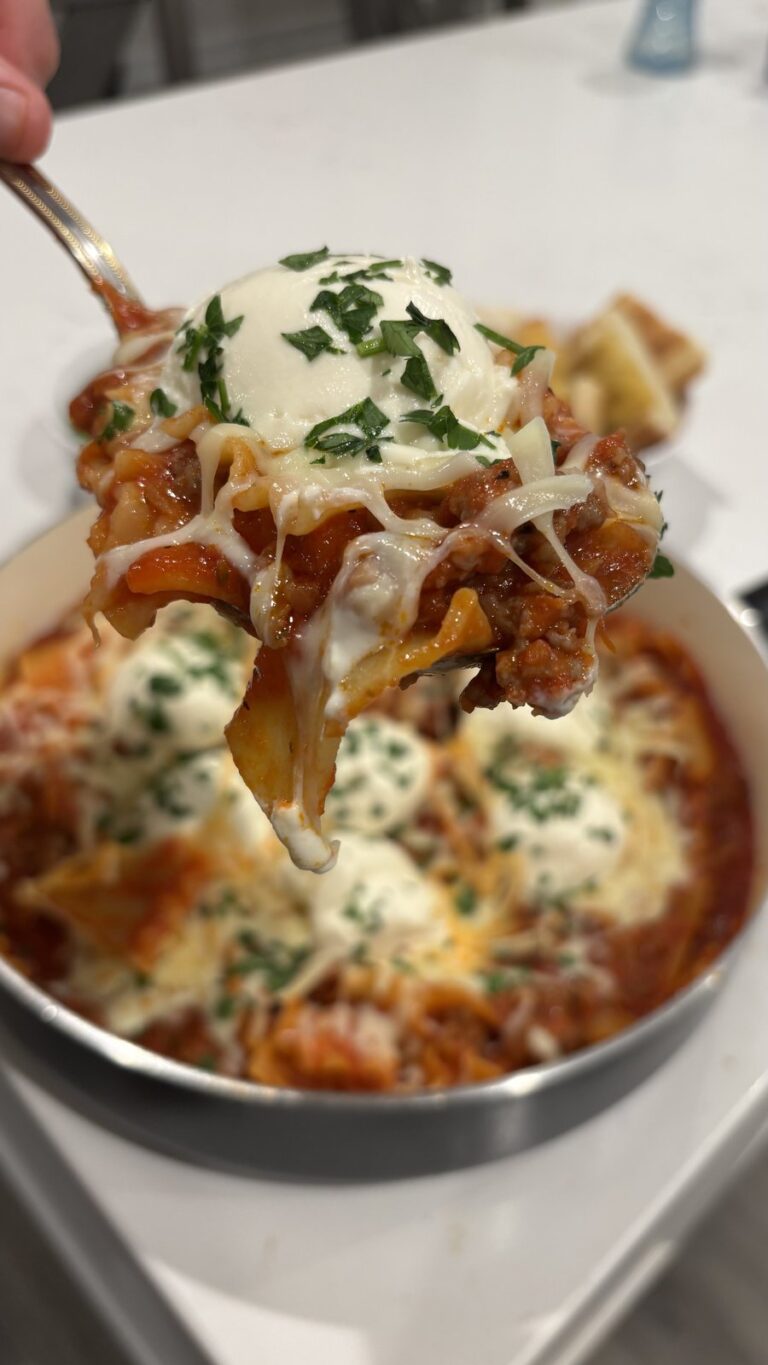 A close-up shot of a forkful of cheesy baked lasagna being lifted from a skillet, garnished with fresh parsley and melted mozzarella.