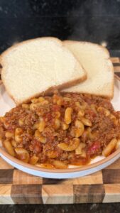 A bowl of macaroni goulash with ground beef and tomato sauce served with two slices of buttered white bread on a wooden cutting board.