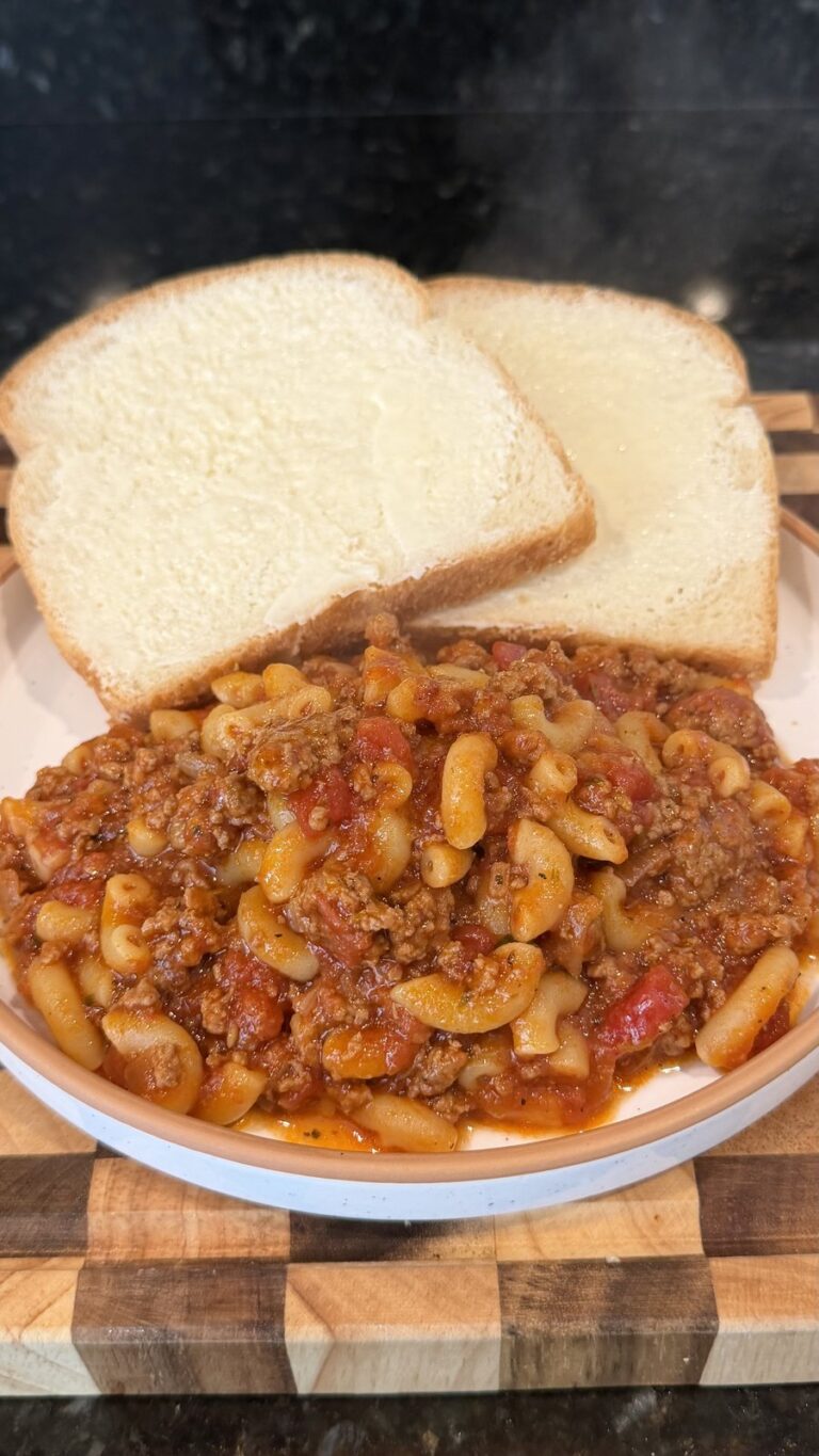 A bowl of macaroni goulash with ground beef and tomato sauce served with two slices of buttered white bread on a wooden cutting board.