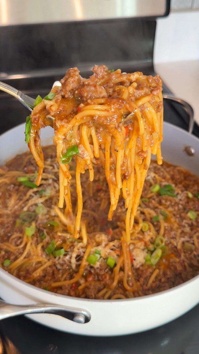 A fork lifts a serving of saucy spaghetti with ground beef and scallions from a white skillet on a stovetop.