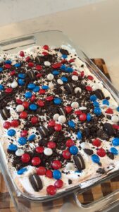 A patriotic-themed no-bake Oreo dessert topped with crushed cookies and red, white, and blue M&M's sits in a glass baking dish on a kitchen counter.