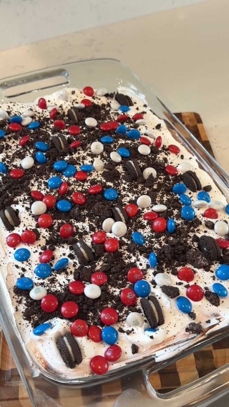 A patriotic-themed no-bake Oreo dessert topped with crushed cookies and red, white, and blue M&M's sits in a glass baking dish on a kitchen counter.