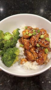 A close-up overhead shot of a white bowl containing glazed sesame chicken with green onions over white rice, served alongside roasted broccoli.