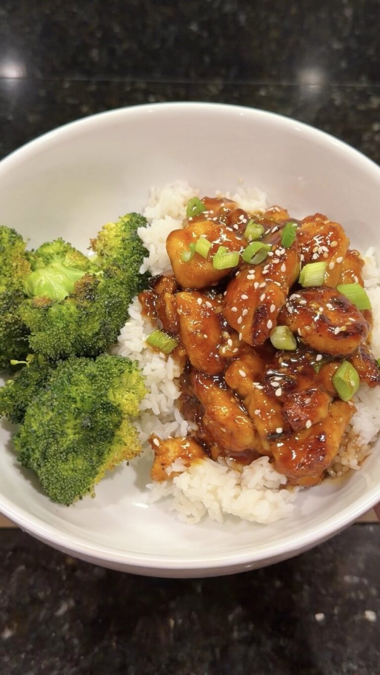 A close-up overhead shot of a white bowl containing glazed sesame chicken with green onions over white rice, served alongside roasted broccoli.
