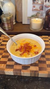 A bowl of creamy cheesy soup topped with shredded cheddar and bacon bits sits on a wooden cutting board with a lit candle in the background.