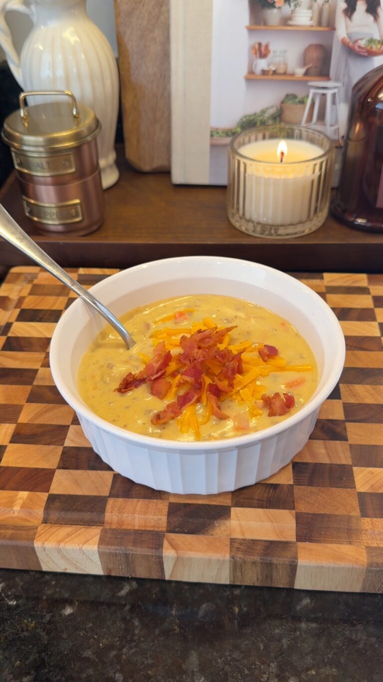 A bowl of creamy cheesy soup topped with shredded cheddar and bacon bits sits on a wooden cutting board with a lit candle in the background.