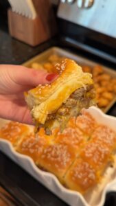 A hand holds up a cheesy beef slider on a Hawaiian roll, pulled apart to show melted cheese and meat, with a tray of sliders and tater tots in the background.