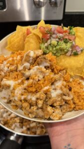 A hand holds a plate of cheesy Mexican rice with chicken, tortilla chips, guacamole, and pico de gallo in a kitchen.