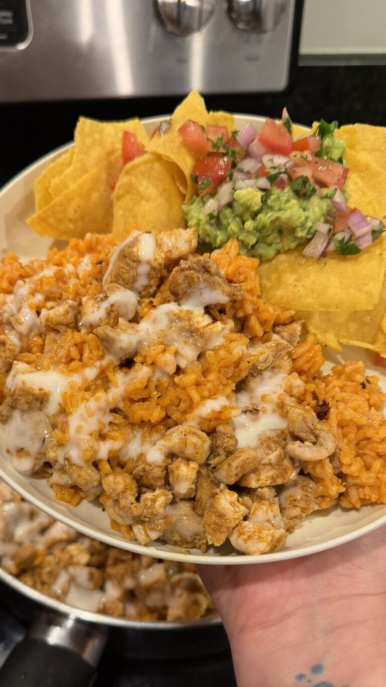 A hand holds a plate of cheesy Mexican rice with chicken, tortilla chips, guacamole, and pico de gallo in a kitchen.