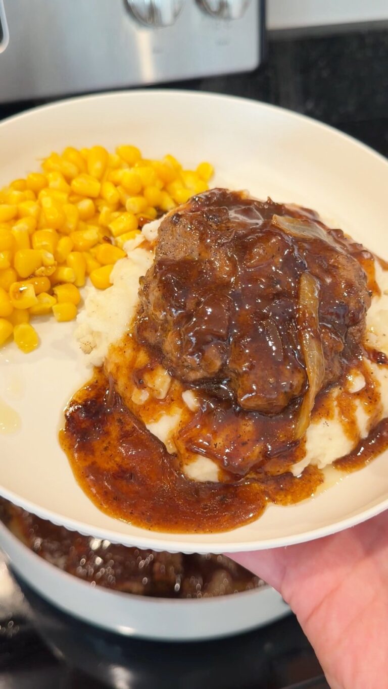 A close-up of a plate with mashed potatoes topped with meat and gravy alongside a serving of corn, held by a hand in a kitchen.